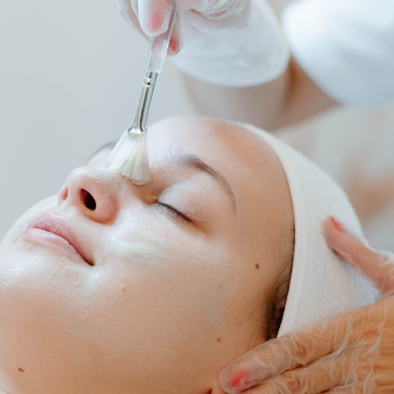 Cosmeticiană aplicând o mască facială cu pensula unei cliente întinse pe pat, în cabinet.